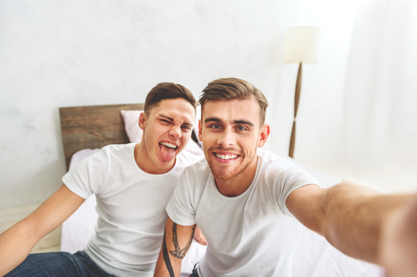 Selfie of gay couple sitting on bed one smiling one sticking his tonge out